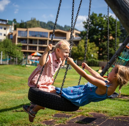 Zwei Mädchen haben Spaß auf der Schaukel am Spielplatz des Kinderhotels in Oberösterreich