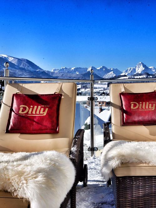 Kuschelige mit Fell bedeckte Liegen auf der sonnigen winterlichen Terrasse des Hotels im Nationalpark. Im Hintergrund die verschneiten Berge